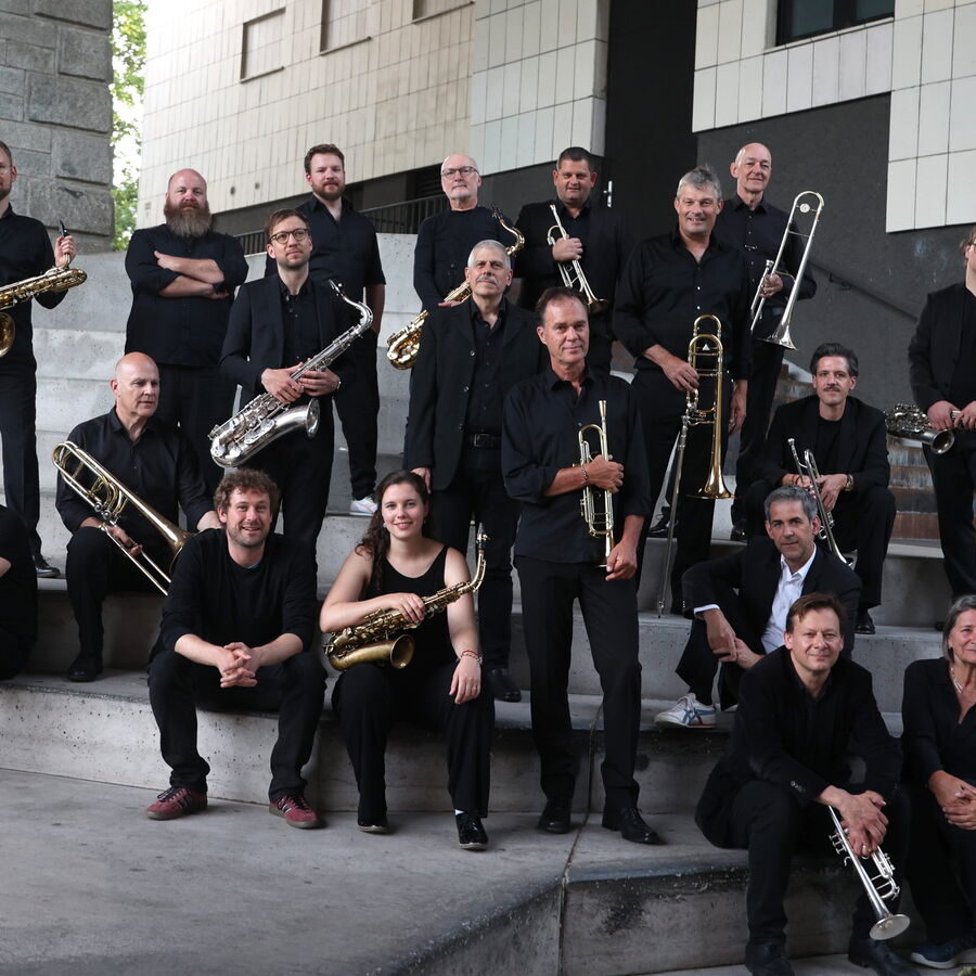 Eine Gruppe von Menschen sitzt und steht auf mehreren Stufen im Außenbereich vor einem modernen Gebäude. Alle tragen dunkle Kleidung und halten verschiedene Blasinstrumente wie Saxophone, Trompeten und Posaunen.
